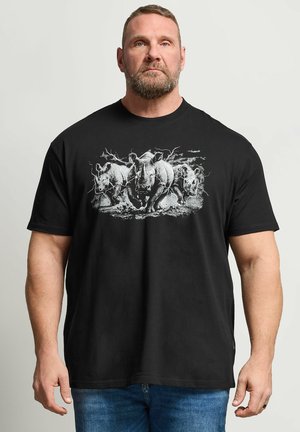 Mężczyzna z krótkimi włosami i brodą, ubrany w czarną koszulkę z grafiką trzech pędzących nosorożców, stoi na tle jednolitego jasnego tła.