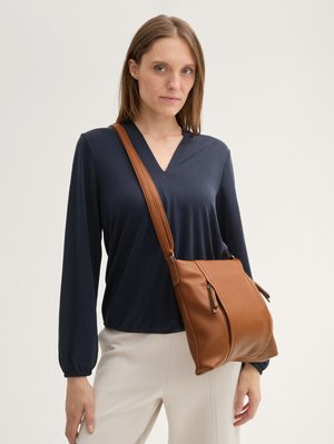 Braun strukturiert, Schultertasche aus Leder mit gewölbtem Design, ausgestattet mit einem Reißverschluss und seitlichen Details. Das Modell trägt ein marineblaues Oberteil und cremefarbene Hosen.