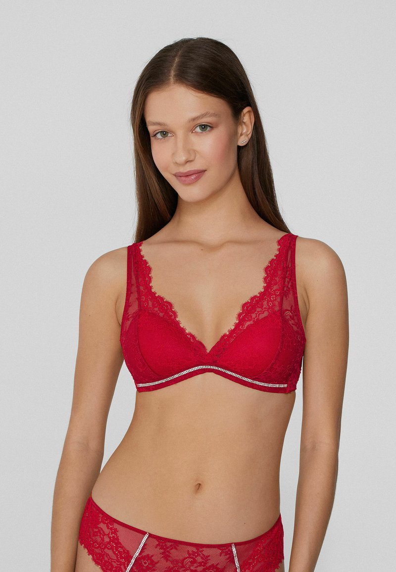 El sujetador de encaje rojo presenta un escote pronunciado, bordes en forma de ondas, tirantes anchos y una banda bajo el busto con detalles plateados.