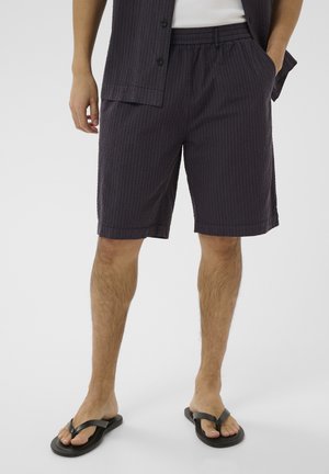 Jambes d'homme portant un short texturé foncé, des tongs noires et une chemise sombre partiellement visible avec des mains dans les poches.