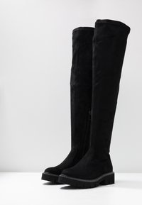 Bottes noires montantes au-dessus du genou en daim doux avec une semelle épaisse, dotées d'une fermeture éclair latérale et de coutures contrastantes le long de la semelle.
