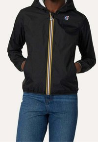 Svart lätt jacka med huva, prydd med en flerfärgad dragkedja och elastiska ärmslut. Bärs över blå jeans, vilket ger en avslappnad passform.