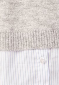 Maglione grigio in lana con orlo a costine, indossato sopra una camicia a righe blu e bianche con un bottone bianco visibile.