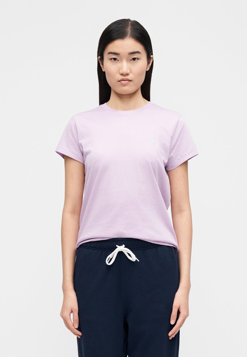 Ung kvinna med rakt svart hår som bär en ljuslila t-shirt och marinblå byxor med vit dragsko, stående mot en enkel ljus bakgrund.