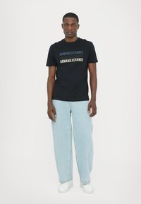 Svart kortermet t-skjorte med "ARMANIEXCHANGE" i varierte farger, sammen med lyseblå løse jeans og hvite sneakers.