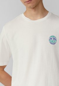 Witte katoenen T-shirt met een ronde afbeelding in groen en blauw met de tekst "ENKELE PLAATS IN HET STADSE UNIVERSE." Korte mouwen, ronde hals.
