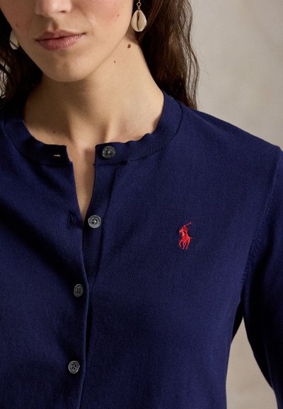 Nő sötétkék gombos kardigánban, piros hímzett póló játékos logóval és fehér kagyló fülbevalóval.