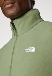 Veste en polaire zippée de couleur verte douce, avec un col haut, un tissu texturé et un logo brodé blanc sur la poitrine gauche.