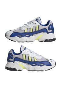 Baskets de sport avec une tige en mesh blanc et gris, des accents bleu marine, des rayures jaunes et une semelle blanche texturée avec un fond noir.