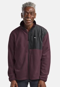 Fleece-Jacke in Bordeauxrot mit einem schwarzen strukturierten Einsatz und einer Reißverschlusstasche. Sie verfügt über einen hohen Kragen und lange Ärmel mit elastischen Bündchen.