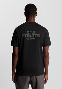Sort bomulds t-shirt med fed hvid tekst på ryggen: "LYLE ATHLETIC" og "FUEL AMBITION." Korte ærmer, rund halsudskæring.