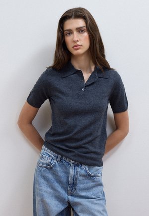 Giovane donna con lunghi capelli castani che indossa una polo scura a maniche corte e jeans azzurri, in piedi contro un muro semplice.