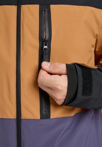 Herrenjacke mit einem Farbblock-Design in orange, schwarz und violett; verfügt über eine Reißverschlusstasche und einen Klettverschluss am Ärmel.