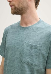 Camiseta de manga corta y cuello redondo en color azul claro, con un bolsillo frontal que presenta un patrón de tela texturizada y ligeramente jaspeada.