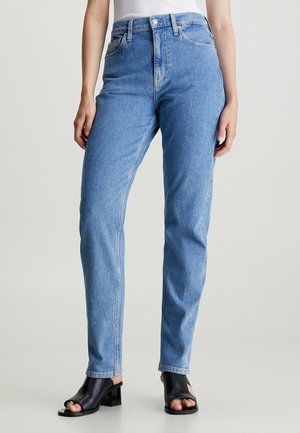 Femme portant un jean taille haute bleu clair et des sandales noires à talons blocs ouverts, debout devant un fond uni.