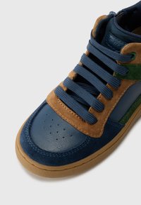Blauwe en bruine high-top sneaker, gemaakt van leren en suède materialen, met een ronde neus, opvallende veters, en een gumkleurige zool met perforaties op de neus.