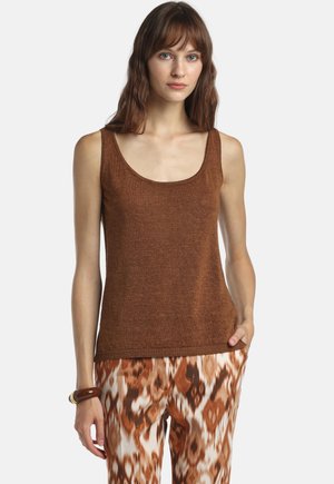 Žena oblečená v hnedom bezrukávovom tričku s výstrihom do U a hnedých nohaviciach s bielo-hnedým vzorom, na ľavom zápästí má náramok.