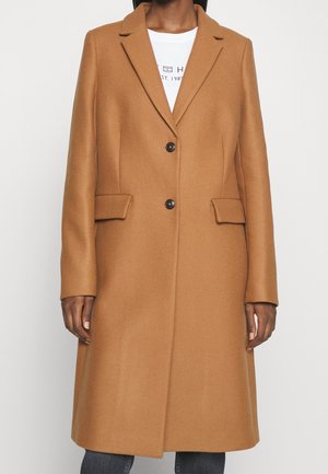 Kvinde iført en lang kamelbrun frakke med notch-revers, to sorte knapper, klaplommer og en hvid skjorte under.