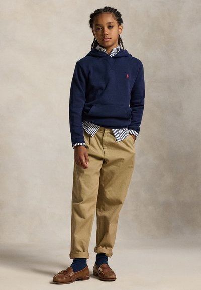 Bernužis su pintomis kasomis stovi apsirengęs tamsiai mėlyną džemperį su kapišonu, dryžuotą marškinių, rudos spalvos kelnes, mėlynas kojines ir rudus mokasinus paprastame fone.