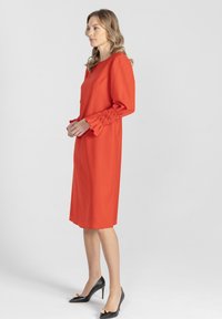 Czerwona sukienka z długimi rękawami, z marszczeniami przy mankietach. Gładki materiał, długość do kolan, zestawiona z czarnymi szpilkami.