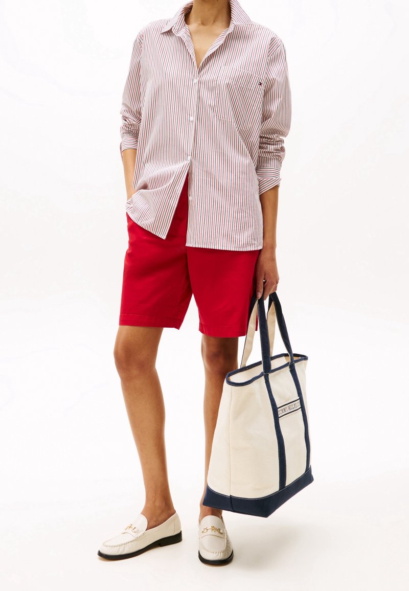 Personne portant une chemise rayée rouge et blanche, un short rouge, des mocassins blancs et tenant un sac fourre-tout blanc avec des poignées et un liseré bleu marine.