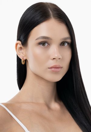 Goldene Creolen mit einem strukturierten, verdrehten Design und einem leichten, polierten Finish. Tragen von einem Model mit glattem, dunklem Haar.
