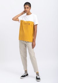 Geel en witte T-shirt met een horizontaal ontwerp, voorzien van een grafische print, gecombineerd met lichtbeige broek en zwarte sneakers.