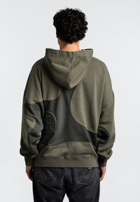 Hanorac verde oliv cu patch-uri texturate, croi supradimensionat și accente întunecate în contrast. Dispune de un logo mare pe spate și o glugă spațioasă.
