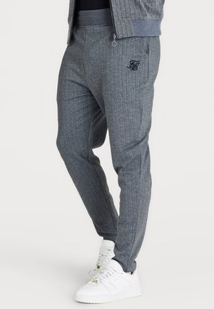 Mężczyzna noszący szare prążkowane spodnie dresowe z czarnym logo na udzie i białe sneakersy, stojący na jednolitym tle.
