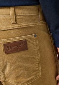 Teddygrabben broek in tan met een textuur, featuring een bruine leren patch met "Wrangler" gepreefd en een zijzak met een metalen stud.