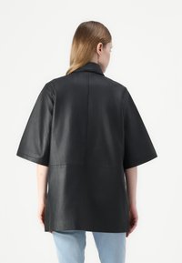 Jachetă scurtă din piele neagră, cu mâneci scurte, guler structurat și detalii pe cusătura din spate, având o textură netedă și un design minimalist.