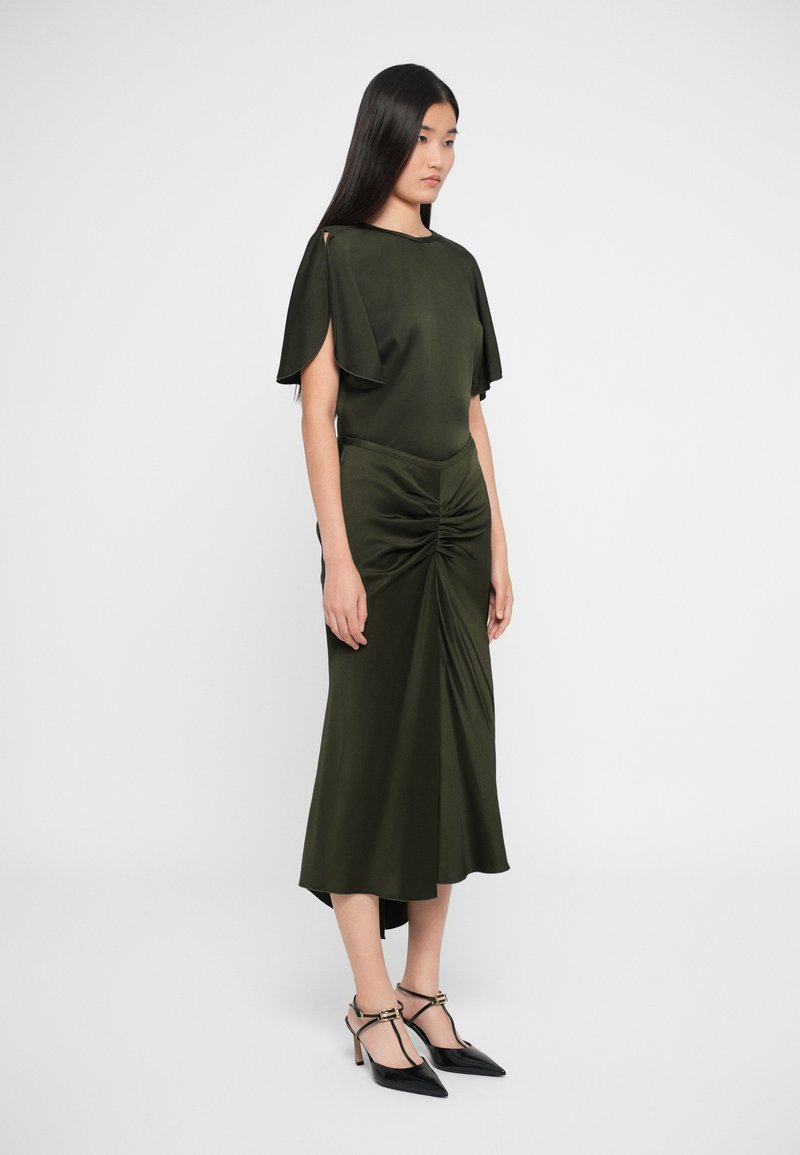 Robe midi vert olive avec des manches flottantes, un design froncé à l'avant et un ourlet asymétrique. Assortie à des escarpins noirs à bouts pointus.