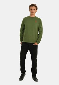 Green sweatshirt with long sleeves, ribbed cuffs and hem, featuring a small red logo. Paired with black pants and black sneakers.