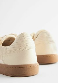Valkoinen sneaker-malli, jossa on teksturoitu nahkainen päällinen, pehmustettu nilkkaosa ja ruskea kumipohja. Huomaamatonommelta ompeluja ja reikiä koristeena.