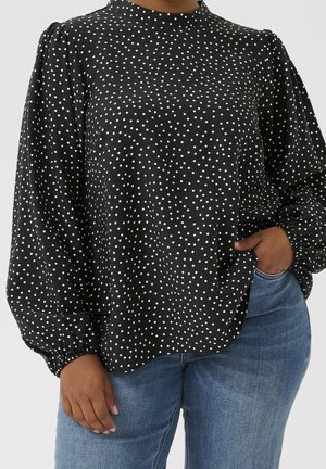Blouse noire à pois blancs, col montant, manches bouffantes et poignets froncés. Associée à un jean bleu clair.