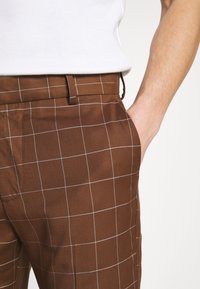Brown plaid trousers feature a smooth texture, with white grid lines and a straight design. The waistband has a belt loop and side pockets.