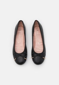 Para czarnych skórzanych balerinek z zaokrąglonymi palcami i małymi kokardkami ze złotymi końcówkami, umieszczona z góry na jednolitym białym tle.