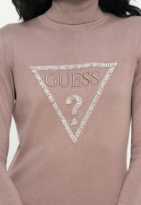 Primo piano di un dolcevita color malva con un logo triangolare GUESS decorato con perline e rhinestones sul petto.