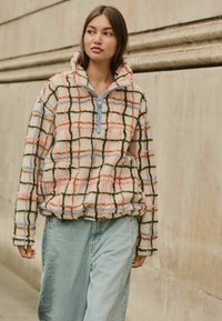 Pulover din fleece cu fermoar, prezentând un model texturat, în carouri multicolore în nuanțe de roz, albastru și verde pe un fundal crem.