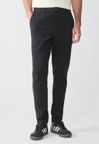 Black chino pants with a slim fit, featuring side pockets and a single button closure. Worn with black sneakers and white stripes.