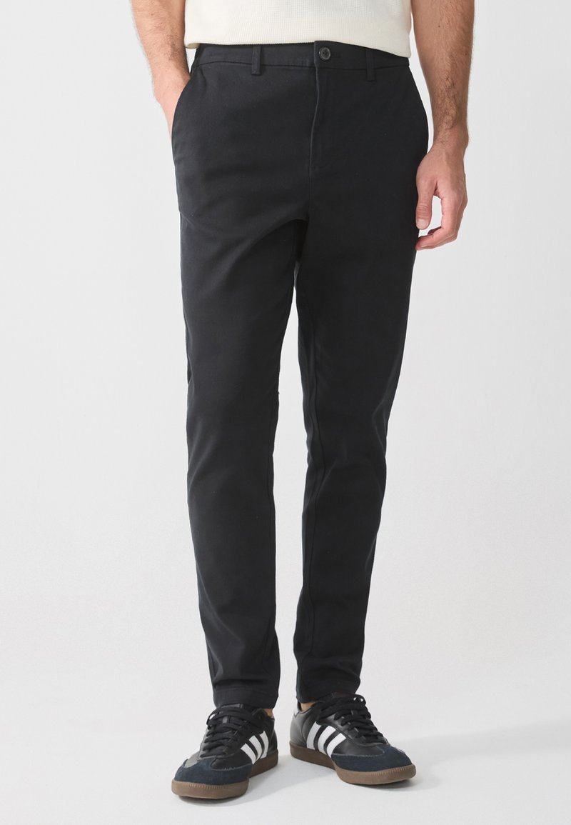 Black chino pants with a slim fit, featuring side pockets and a single button closure. Worn with black sneakers and white stripes.