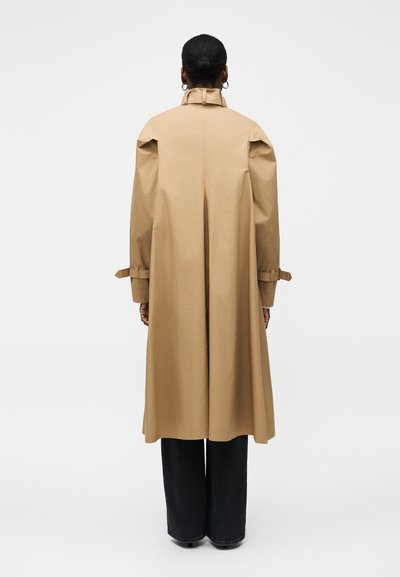 Femme avec les cheveux en chignon porte un long trench-coat beige avec des détails de ceinture sur les manches, debout de dos sur fond blanc.
