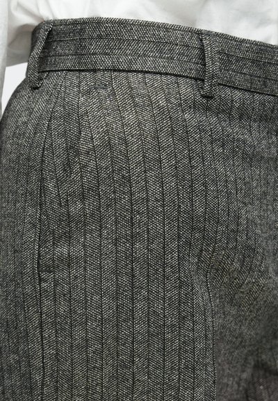 Krupan plan siva herringbone uzorka vune hlača nosi se s ubodenom bijelom košuljom, naglašavajući pojas i petlje za remen.