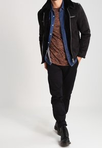 Schwarze Wildlederjacke mit einem Kunstfellkragen, über einem braun-grau gestreiften Hemd und dunklen Hosen geschichtet, vervollständigt mit schwarzen Schnürstiefeln.