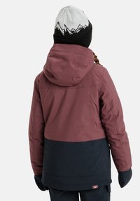 Bordeauxrode en marineblauwe waterdichte jas met kap, met een gladde textuur en een logo patch aan de zijkant. Draag ik met een zwarte gebreide muts.