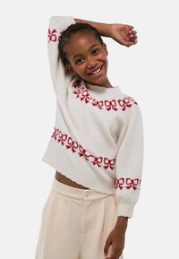 Pull blanc avec motif à ruban rouge, coupe courte, manches larges et ourlet côtelé. Texture douce, associé à un pantalon beige décontracté.