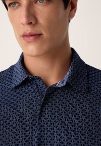 Tengerészkék ing geometriai mintával, begombolt gallérral és világos kontrasztos részlettel a belső galléron. Simább anyag textúra.