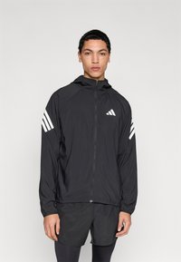 Zwarte lichtgewicht jas met ritssluiting aan de voorkant, capuchon en drie witte strepen op elke mouw. De stof heeft een gladde, matte textuur.