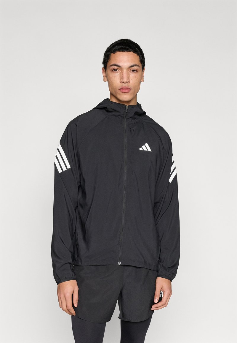 Zwarte lichtgewicht jas met ritssluiting aan de voorkant, capuchon en drie witte strepen op elke mouw. De stof heeft een gladde, matte textuur.