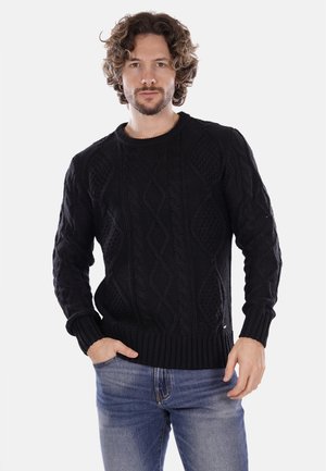 Maglione nero lavorato a maglia con motivi a trecce testurizzati, scollo a girocollo e polsini e vita a coste, abbinato a jeans blu.